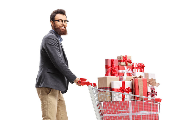 Bearded man pushing a shopping cart full of presents and smiling at the camera isolated on white background - Foto, afbeelding