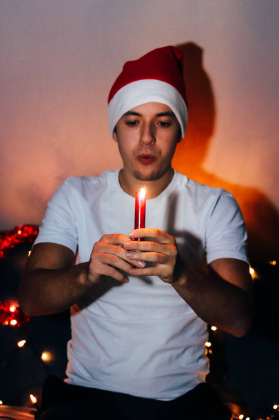 Mladý muž se chystá sfouknout svíčku na vánočním večírku a přeje si něco na nový rok2021. Vánoční klobouk. Koncept klidu a pohody během vánočních svátků. - Fotografie, Obrázek