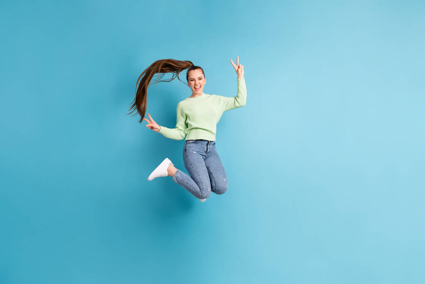 Фотография прыгающей девушки с длинными волосами в полный рост, на которой изображены V-образные обе руки, изолированные на ярко-голубом фоне - Фото, изображение