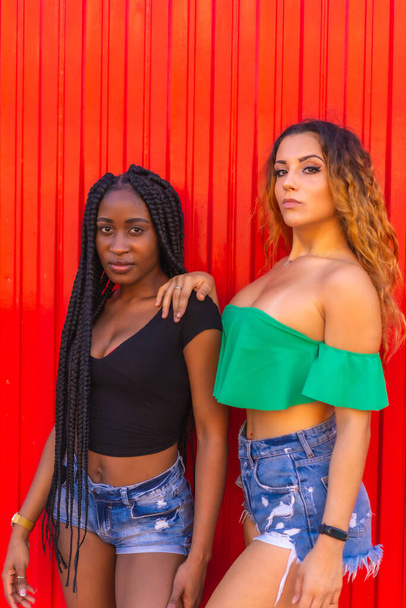 Lifestyle of friends, Caucasian blonde girl and black girl with big braid on a red background. Modern photography of urban girls in the city - Foto, afbeelding