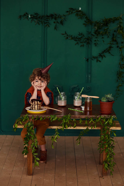 Cute curly little boy celebrates his birthday with delicious holiday pancakes. Child in a conical hat celebrates at a home party in a stylish room with green walls decorated with leaves. - Valokuva, kuva