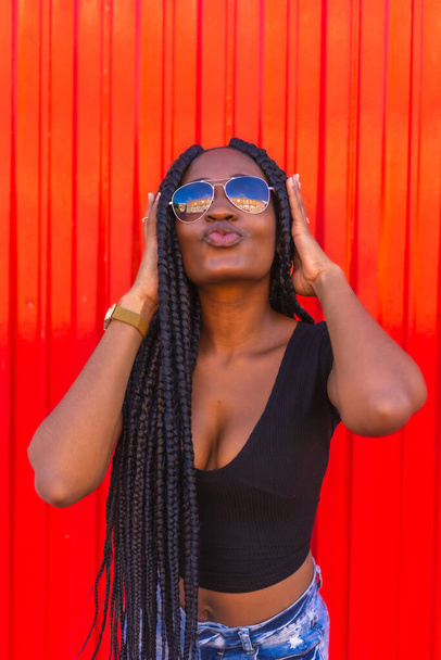Lifestyle, black girl with big braid in black t-shirt, denim shorts and sunglasses on a red background. Portrait of a young trap dancer - Фото, изображение