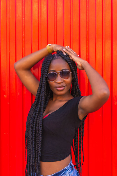 Lifestyle, black girl with big braid in black t-shirt, denim shorts and sunglasses on a red background. College girl enjoying summer days - Zdjęcie, obraz