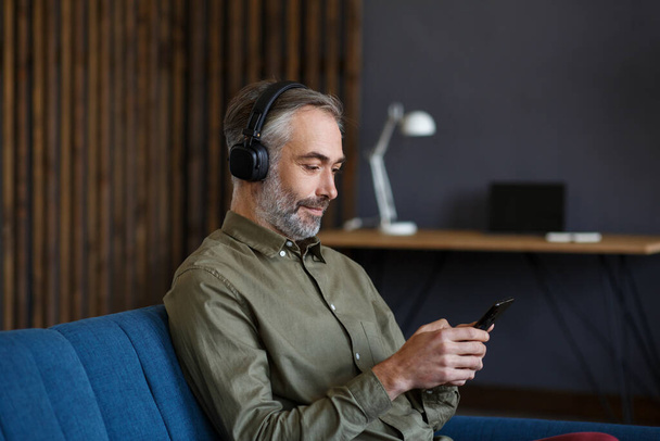 Senior man in headphones listening music on smart phone using music app. Portrait of handsome grey-haired man in earphones and mobile phone at home. Relaxation, leisure. Enjoy listening to music - Foto, Bild