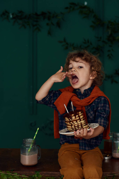 Cute curly little boy celebrates his birthday with delicious holiday pancakes. Child in a conical hat celebrates at a home party in a stylish room with green walls decorated with leaves. - Foto, immagini