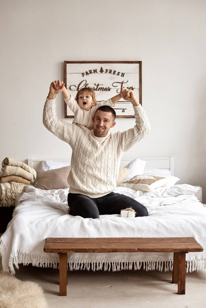 Christmas time. Dad and his little daughter have fun and play sitting on the bed in a bright bedroom with Christmas decor. Concept of a happy childhood and fatherhood. - Фото, зображення