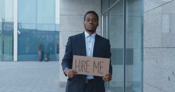 Triste manager afroamericano in cerca di lavoro, a piedi vicino al centro commerciale con poster HIRE ME, slow motion - Filmati, video