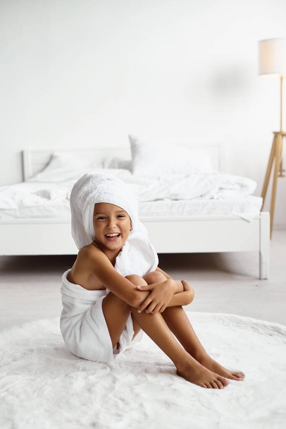 Portrait of a cute little girl with hair wrapped in a bath towel after a bath or shower. Child has fun in a bright hotel room. Spa procedure, bath textile. - Foto, Imagem