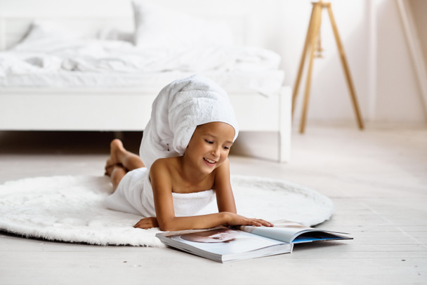 Little model girl wrapped in a bath towel lying on the floor on a white carpet and reading a fashion magazine. Child poses in a bright hotel room or bedroom at home. Concept of a carefree childhood. - Fotoğraf, Görsel