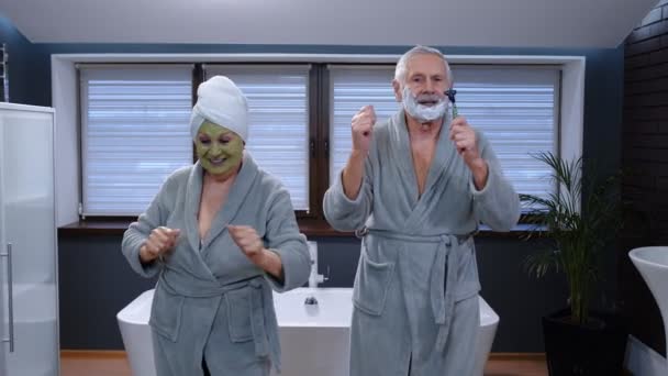 Ältere Großmutter mit grüner Gesichtsmaske und Großvater mit Rasierschaum tanzen im Badezimmer - Filmmaterial, Video