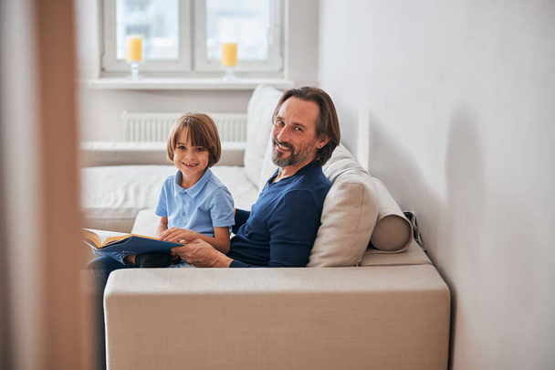 Очаровательный ребенок и его бородатый отец отдыхают на диване - Фото, изображение