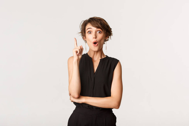 Portrait de femme séduisante excitée en tenue formelle levant le doigt dans geste eureka, avoir une idée, fond blanc - Photo, image