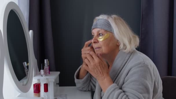 Ηλικιωμένη ηλικιωμένη γιαγιά που βάζει αντιρυτιδικό κάλυμμα στο μάτι και μακιγιάρεται στο σπίτι - Πλάνα, βίντεο