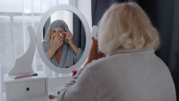 Vanha vanhempi nainen isoäiti hoitaa ihoa silmien ja ryppyjen lähellä, meikkaamalla kotona - Materiaali, video
