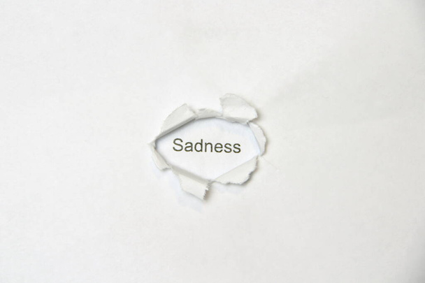 Word Sadness on white isolated background, the inscription through the wound hole in paper. Concept of gender identity and sexual minorities. Stock photo for web and print with empty space for text - Photo, image