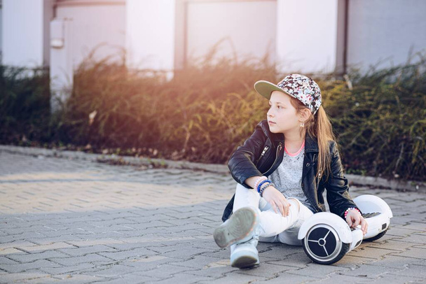 10-letnia dziewczyna w czapce bejsbolowej siedząca na chodniku z deską. - Zdjęcie, obraz