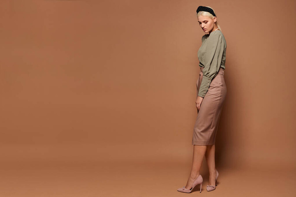 Full-length studio shot of a model girl with blond hair wearing midi size skirt and blouse isolated at the beige background, side view - Фото, зображення
