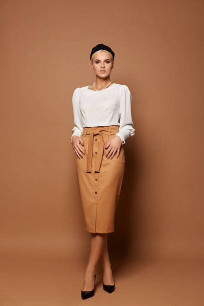Elegant young woman with blond hair in midi size skirt and white blouse posing at the beige background, studio shot - Foto, Imagem