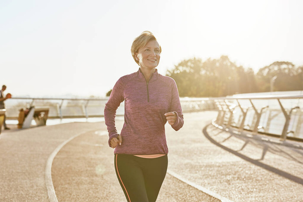 Βελτίωση της υγείας. Πορτρέτο της ευτυχούς κίνητρα ώριμη γυναίκα σε αθλητικά ρούχα τρέχει σε εξωτερικούς χώρους σε μια ηλιόλουστη μέρα - Φωτογραφία, εικόνα
