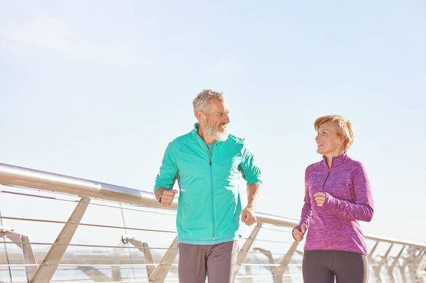 Uaktywnij swoje życie. Aktywna dojrzała para rodzinna w odzieży sportowej uśmiechająca się do siebie podczas biegania razem w słoneczny poranek. Radosna para seniorów ćwiczących na świeżym powietrzu - Zdjęcie, obraz