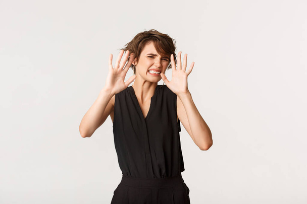 Disgusted young woman grimacing from cringe content, trying to cover eyes with hands, see embarrassing thing, standing white background - Photo, Image