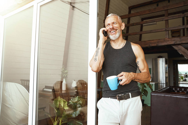 Hyviä uutisia. Onnellinen keski-ikäinen valkoihoinen mies puhuu matkapuhelimella juodessaan kahvia lähellä modernin asuntonsa parveketta. - Valokuva, kuva