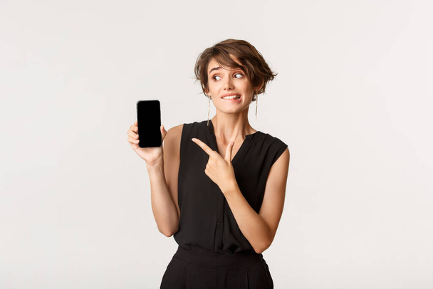 niezdecydowana słodkie dziewczyna wskazując palcem na ekran smartfona i gryzienie wargi wahają, stojąc biały tło - Zdjęcie, obraz