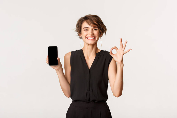 Satisfied young happy woman showing mobile phone screen and okay gesture, guarantee or recommend something, standing white background - Fotó, kép