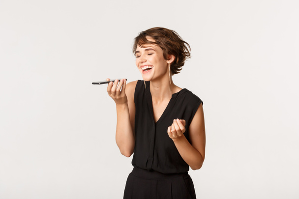 Chica hermosa despreocupada riendo mientras graba el mensaje de voz en el teléfono móvil, de pie fondo blanco - Foto, Imagen