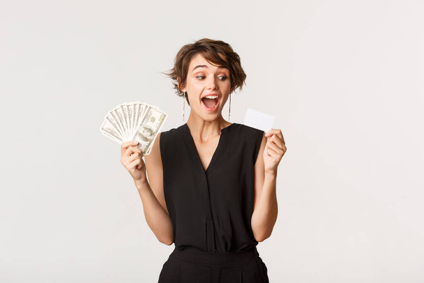 Amazed elegant woman looking at credit card, holding money, standing white background - Foto, immagini