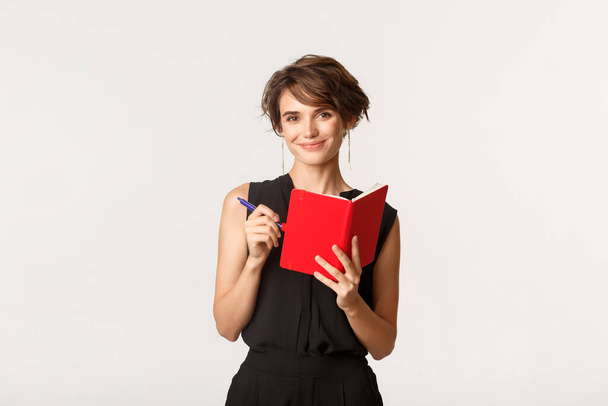 Lächelnde attraktive Unternehmerin, die ihren Terminkalender in Planer schreibt, Notizbuch und Stift in der Hand, vor weißem Hintergrund stehend - Foto, Bild