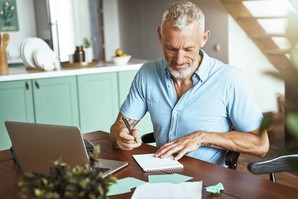 Επικεντρωμένος μεσήλικας καυκάσιος άντρας που κρατάει σημειώσεις ενώ κάθεται στο τραπέζι στο σπίτι κατά τη διάρκεια της απομακρυσμένης εργάσιμης ημέρας - Φωτογραφία, εικόνα