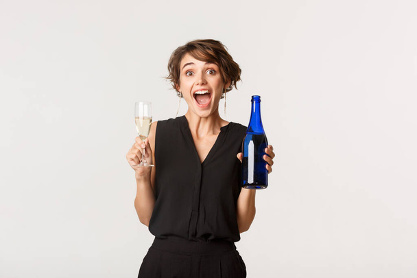 Радостная женщина наслаждается вечеринкой, держа бутылку и бокал шампанского, стоя на белом фоне - Фото, изображение