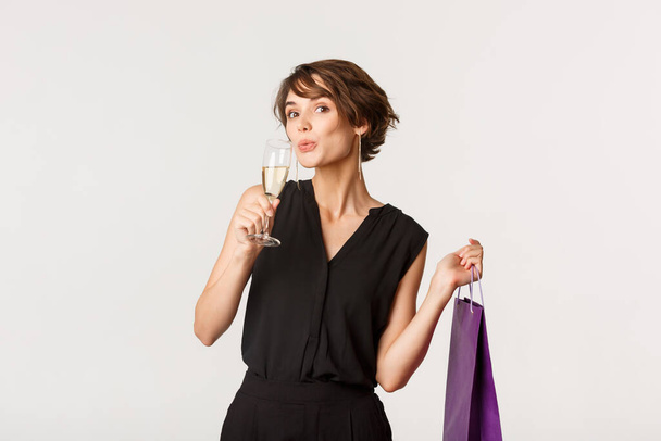 Attractive fashionable woman sipping champagne and holding gift box on formal party, standing over white background - Photo, Image
