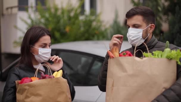 Les jeunes couples masqués tiennent un emballage écologique avec des aliments frais après les achats au supermarché pendant la quarantaine. Homme avec sa femme par le masque de décollage à la maison - Séquence, vidéo
