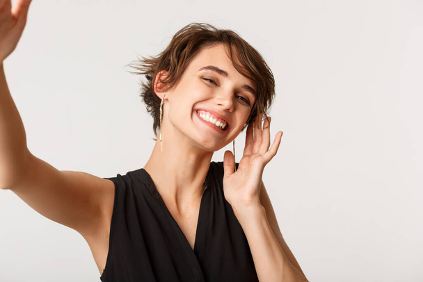 Bild einer eleganten Frau, die ein Selfie mit dem Smartphone macht, sorglos lächelt und vor weißem Hintergrund steht - Foto, Bild