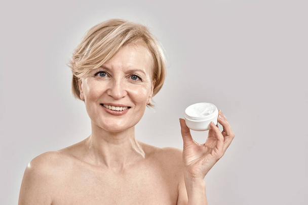 Portrait of beautiful middle aged woman smiling at camera, holding moisturizing facial cream while posing isolated over grey background - Fotografie, Obrázek