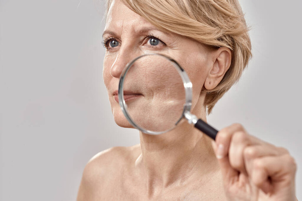 Primer plano retrato de una atractiva mujer de mediana edad mirando a un lado, sosteniendo una lupa y mostrando sus arrugas, posando aislada sobre un fondo gris - Foto, imagen