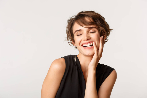 Κοντινό πλάνο της κομψής στιλάτης γυναίκας κλείστε τα μάτια και γελώντας coquettish, στέκεται πάνω από λευκό φόντο - Φωτογραφία, εικόνα