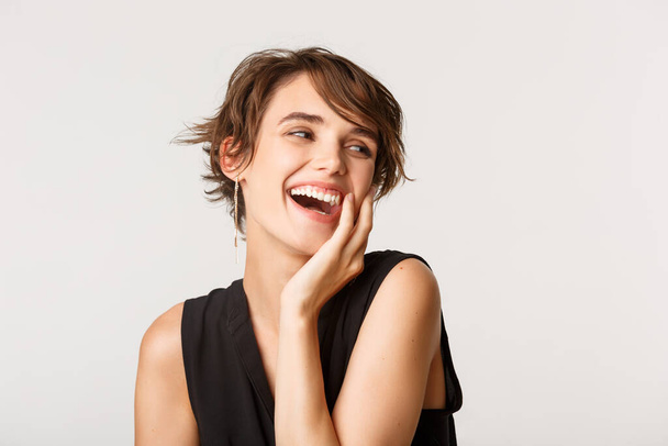 Close-up of elegant stylish woman looking to the right, laughing and gently touching face, standing over white background - Foto, afbeelding