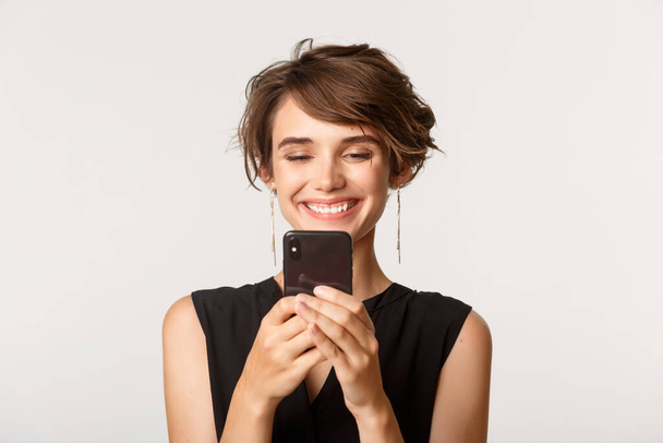 Lähikuva tyytyväinen nuori nainen katselee matkapuhelinta tyytyväinen hymy, seisoo valkoisella taustalla - Valokuva, kuva