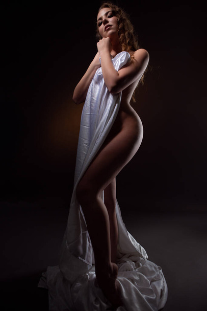 a beautiful naked woman stands modestly covering her magnificent body with a white cloth. Photo on a dark background - Фото, зображення