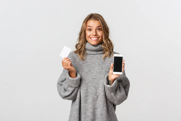 Pretty blond girl in grey sweater, showing smartphone screen with credit card for shopping online, standing against white background - Foto, Imagem