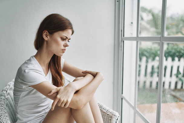Mujer triste cerca de la ventana abraza sus piernas y se sienta  - Foto, Imagen