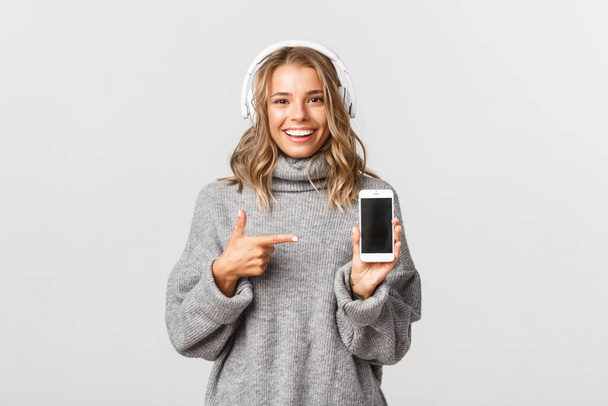 Studio shot of attractive blond girl in grey sweater, listening music in wireless headphones, showing smartphone screen and smiling, white background - Photo, Image