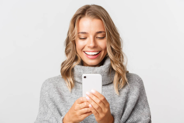 zbliżenie atrakcyjnej blondynki w szarym swetrze, sprawdzanie wiadomości przez telefon lub zakupy online, patrzenie na smartfona ze szczęśliwą twarzą, stanie nad białym tłem - Zdjęcie, obraz