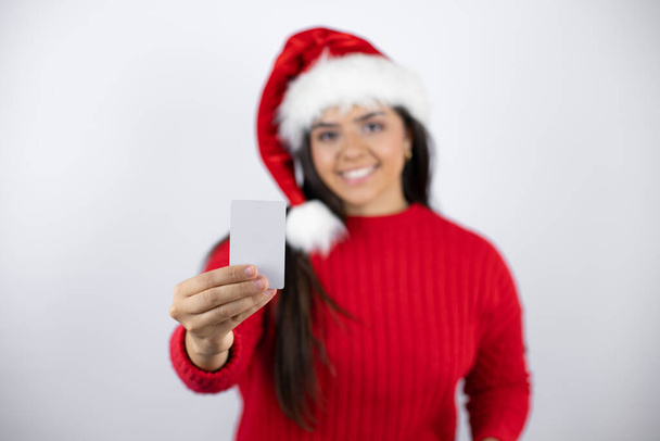 Beyaz arka planda Noel Baba şapkası takan güzel bir kadın gülümsüyor ve elinde beyaz bir kart tutuyor. - Fotoğraf, Görsel