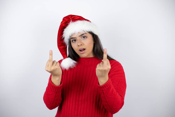 junge schöne Frau trägt einen Weihnachtsmannhut über weißem Hintergrund und zeigt den Mittelfinger dabei, wie sie dich fickt, schlechte Miene, Provokation und unhöfliche Haltung. Kreischen erregt - Foto, Bild