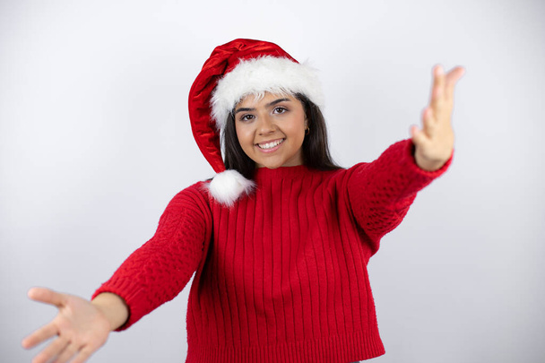 Молода красива жінка в капелюсі Санта над білим тлом дивиться на камеру, посміхаючись з відкритими руками для обіймів
 - Фото, зображення