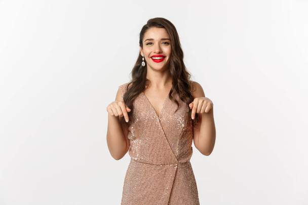 Szczęśliwa piękna kobieta z czerwonymi ustami, uśmiechnięta podczas pokazu promo, ubrana w elegancką sukienkę na przyjęcie świąteczne, wytykająca palcami w dół, stojąca nad białym tłem - Zdjęcie, obraz